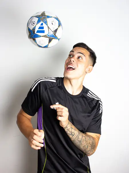 Dudu Footz sorrindo com uma bola de futebol suspensa acima da cabeça, em estúdio