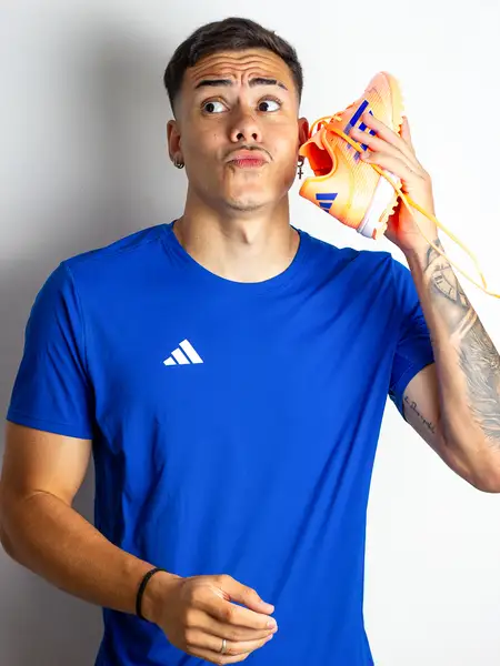 Dudu Footz com uma chuteira encostada na orelha, fazendo cara de quem está ouvindo algo engraçado